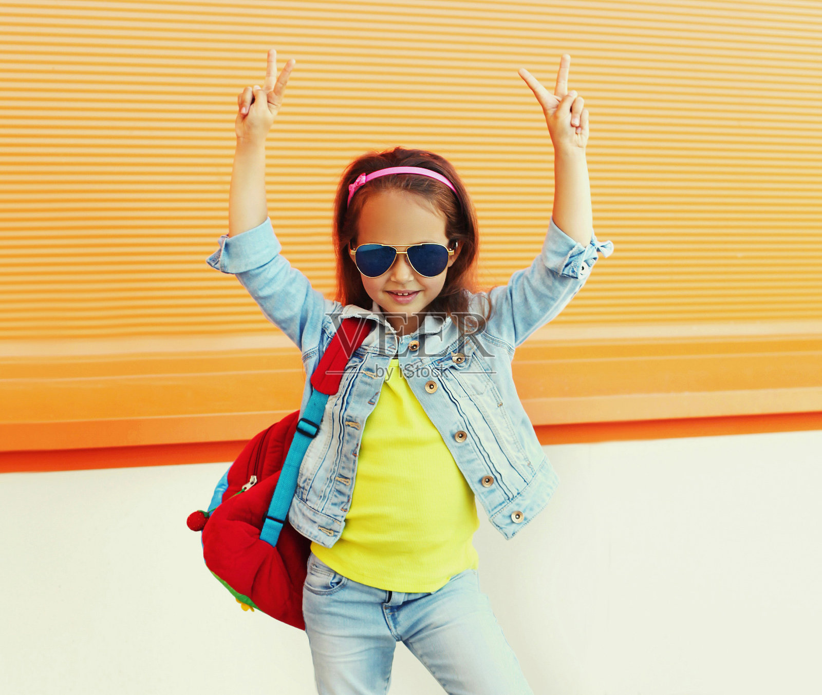 一个小女孩的肖像孩子戴着背包，太阳镜，牛仔裤夹克在城市街道上橙色的背景照片摄影图片