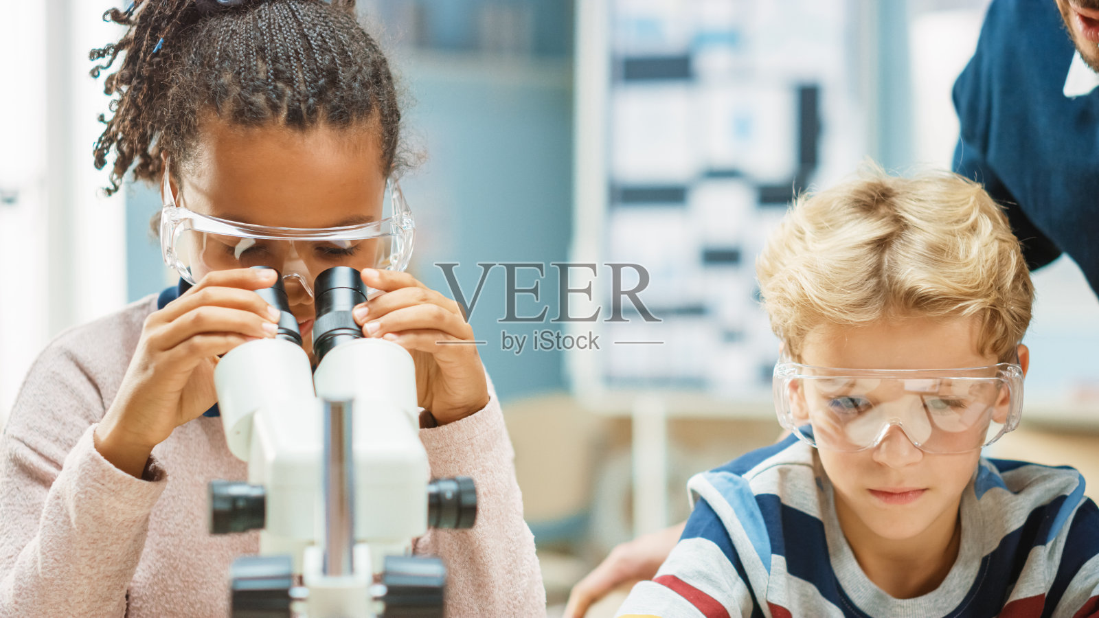 小学科学课堂:可爱的小女孩在显微镜下看，男孩用平板电脑在网上查信息。老师从后面观察照片摄影图片
