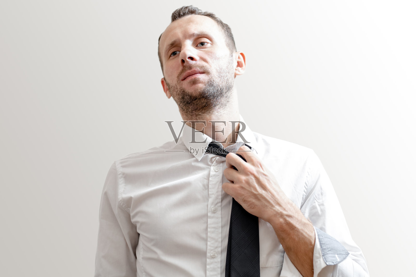 疲倦的年轻人在白色衬衫脱下他的领带在灰色的背景照片摄影图片