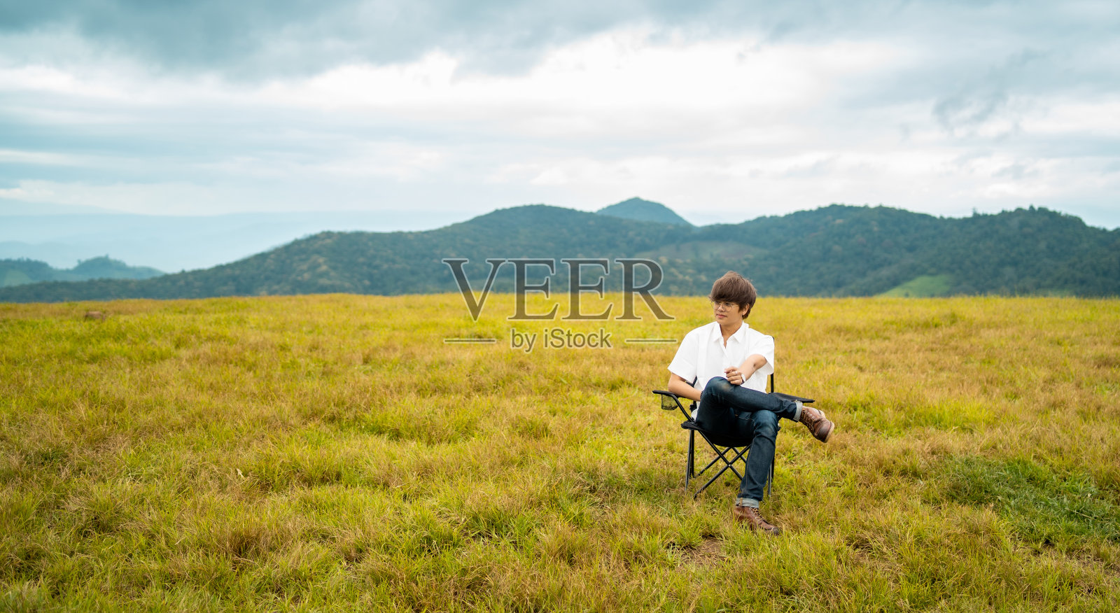 快乐的年轻亚洲人独自坐在户外的椅子上放松在美丽的自然山草地在秋天的一天。寂寞的帅哥享受与户外的生活方式度假照片摄影图片