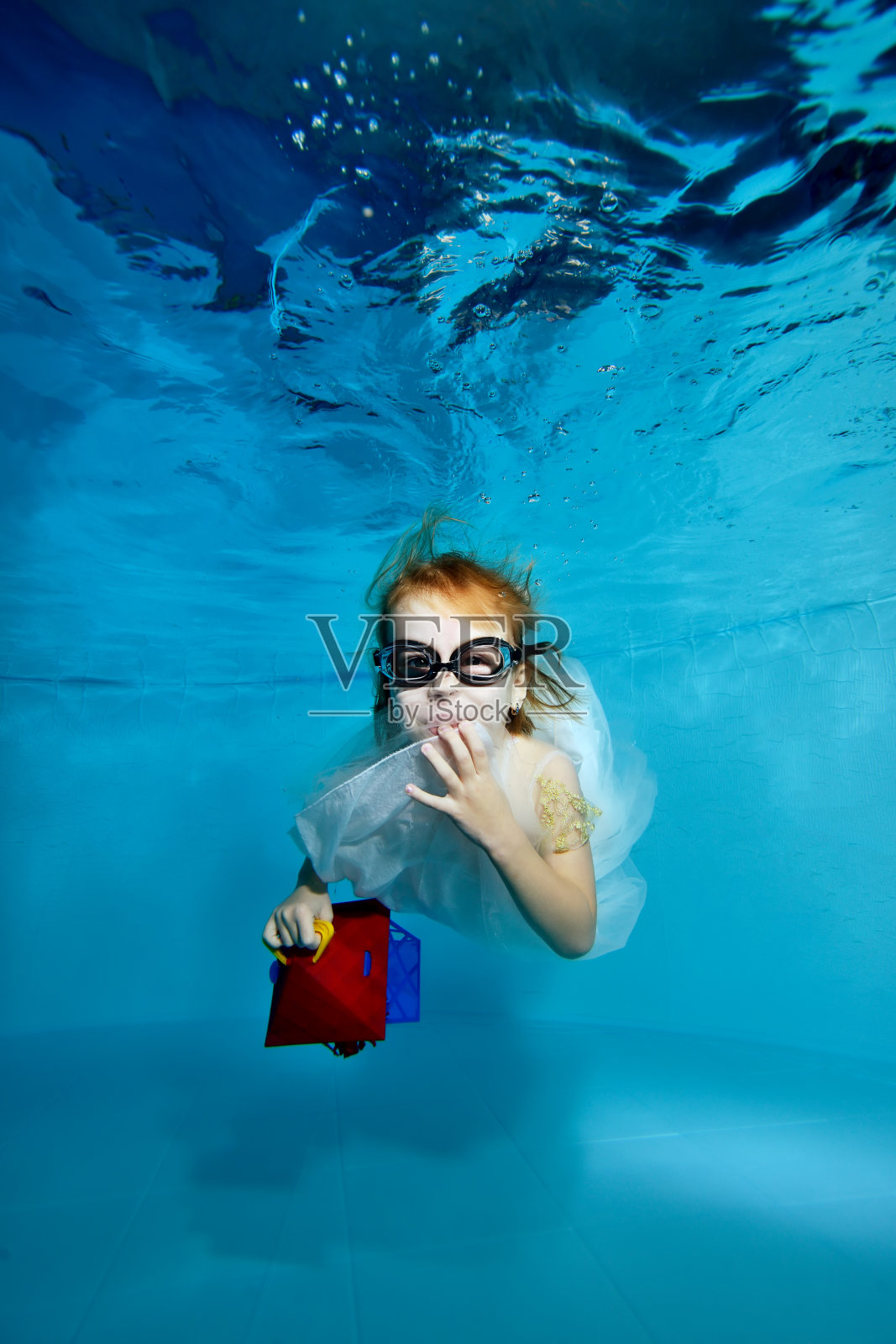 一个有趣的小女孩，穿着蓝色背景的白色裙子，在游泳池的水里游泳，并与一个孩子的礼物摆姿势。她看着镜头，甜甜地笑了。肖像。俯视图照片摄影图片