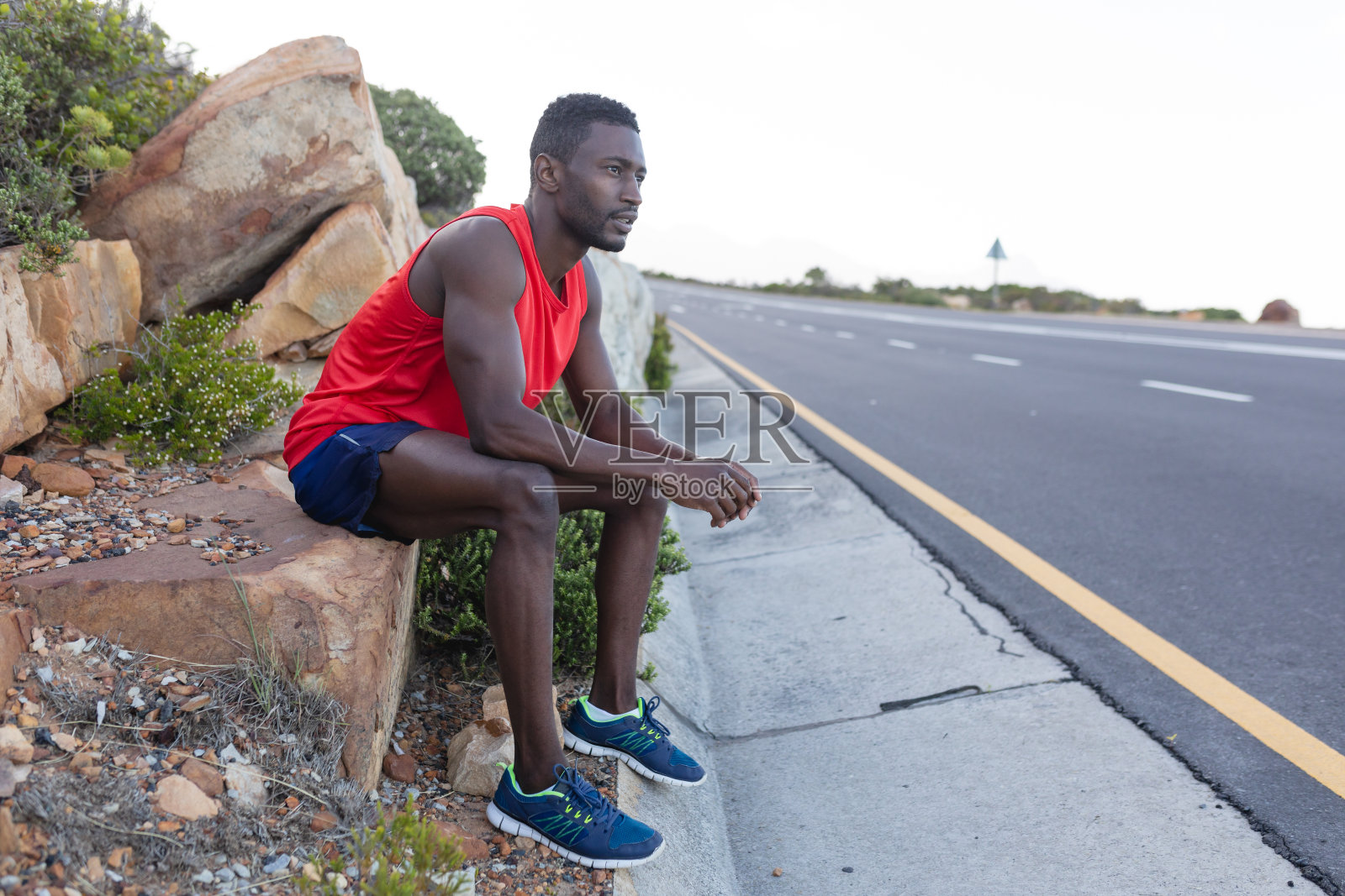穿着运动服的非裔美国人坐在海边公路旁的岩石上照片摄影图片