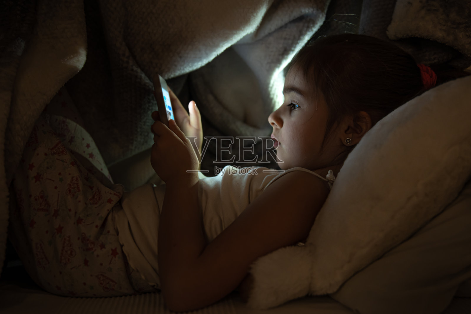 可爱的小女孩晚上在床上玩手机，而不是近距离睡觉。照片摄影图片