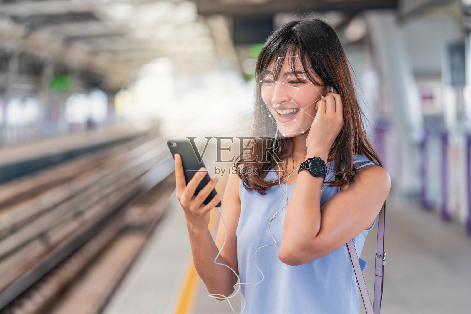 亚洲女性通过智能手机进行人脸识别，在火车站站台听音乐，生物识别验证和人工智能概念照片摄影图片