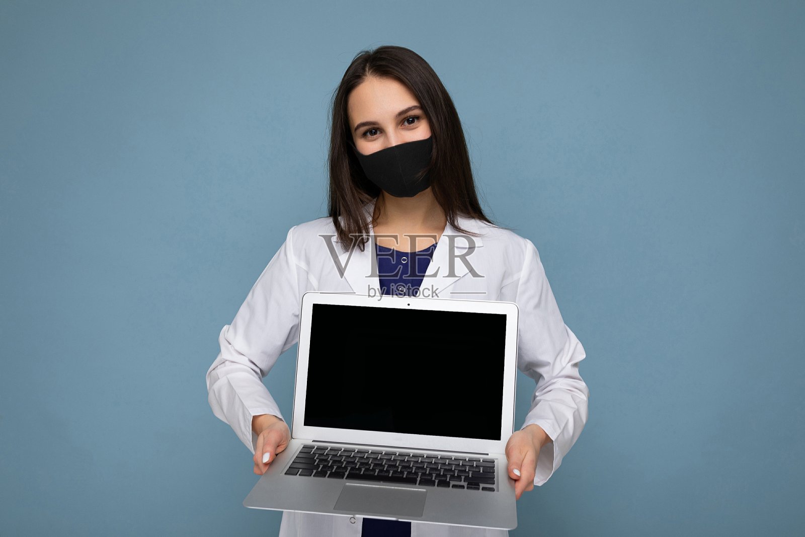 年轻的深色皮肤的女人穿着医学白大褂和黑色面具拿着笔记本电脑，看着孤立的蓝色背景的相机照片摄影图片