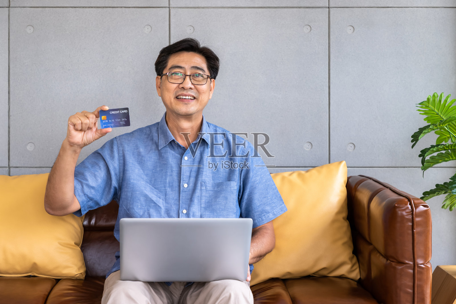 亚裔男子坐在沙发上用手提电脑拿着信用卡进行网上购物和支付，看着摄像头照片摄影图片