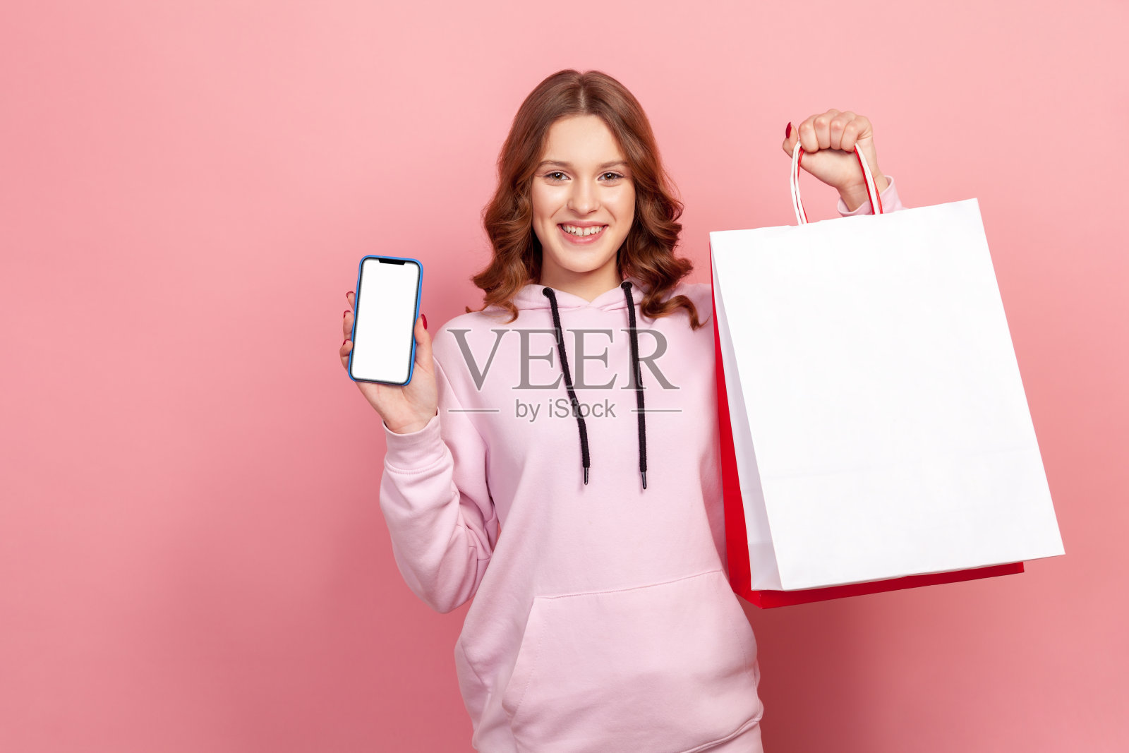 照片上是一个穿着连帽衫的年轻女孩，微笑着拿着纸袋和空屏幕的智能手机，网上购物和现金返还服务照片摄影图片