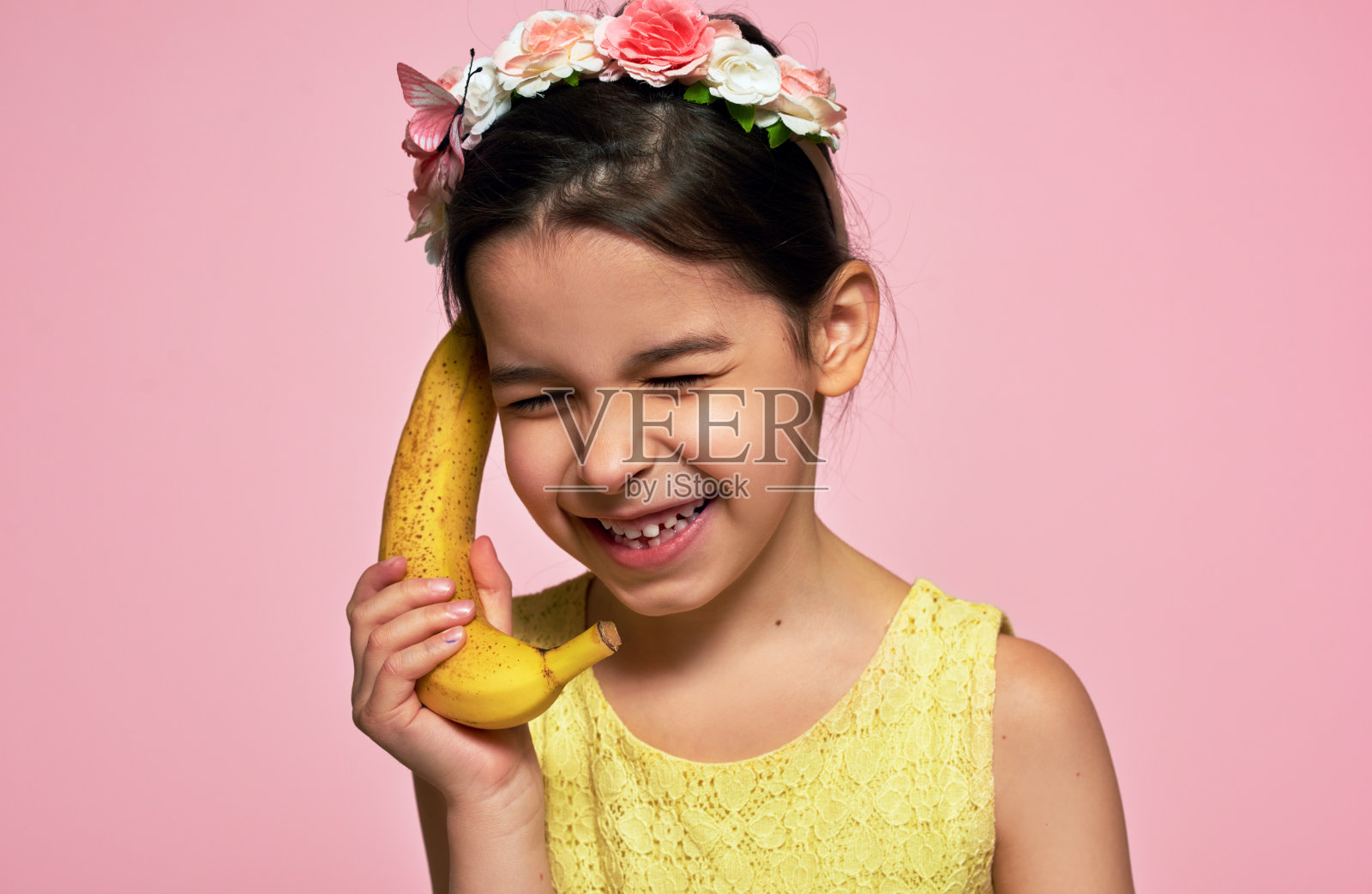 一个快乐的小女孩的特写，笑着拿着一个香蕉，像一个手机，孤立在粉红色的背景。一个穿着黄色连衣裙的快乐的孩子假装对着香蕉说话，就像在玩手机。照片摄影图片