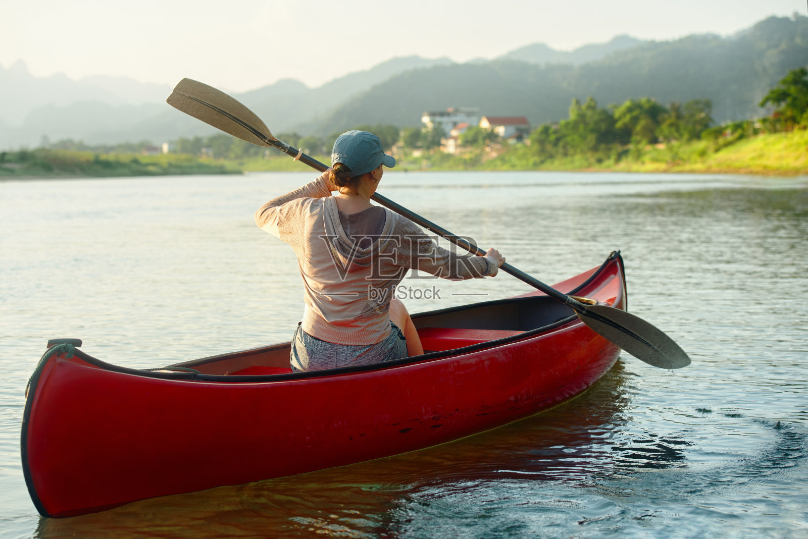 妇女划着桨，在河上漂浮着皮艇，背景是乡村的房屋。照片摄影图片