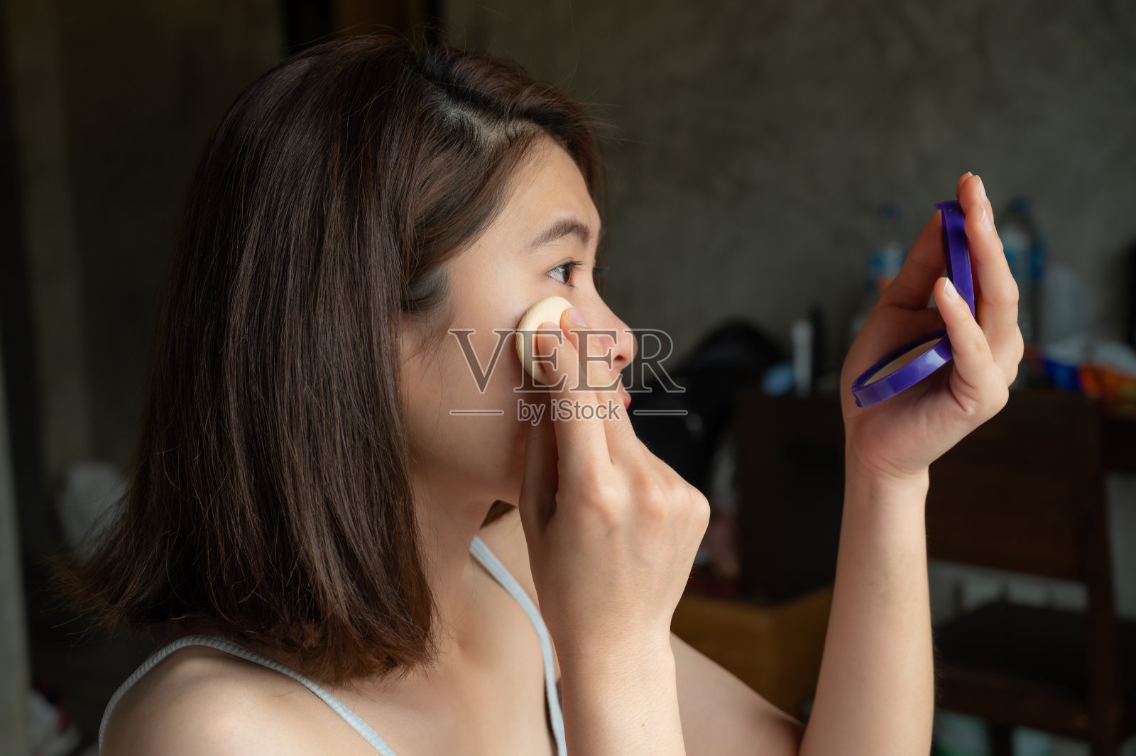 侧面的年轻亚洲妇女将粉底粉扑在她的脸上掩盖瑕疵和改变自然肤色。照片摄影图片