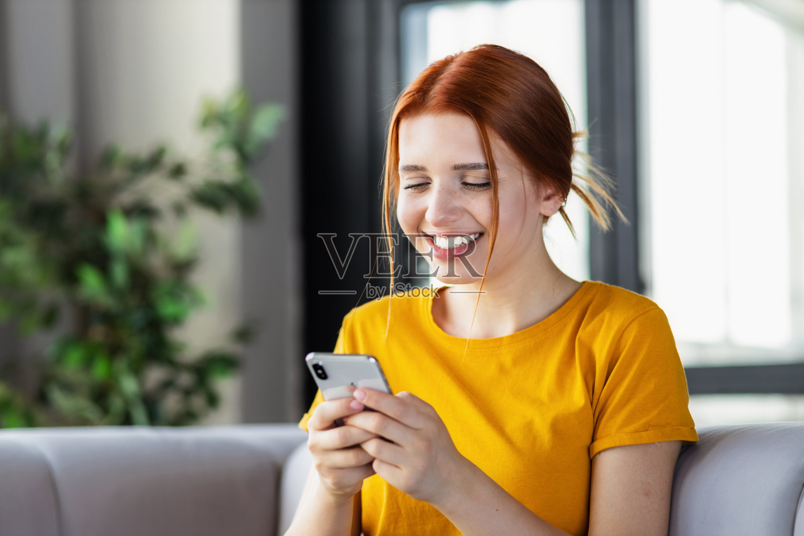 快乐的红发小女孩用手机在线交流，坐在家里的沙发上看搞笑视频，社交网络，微笑着照片摄影图片