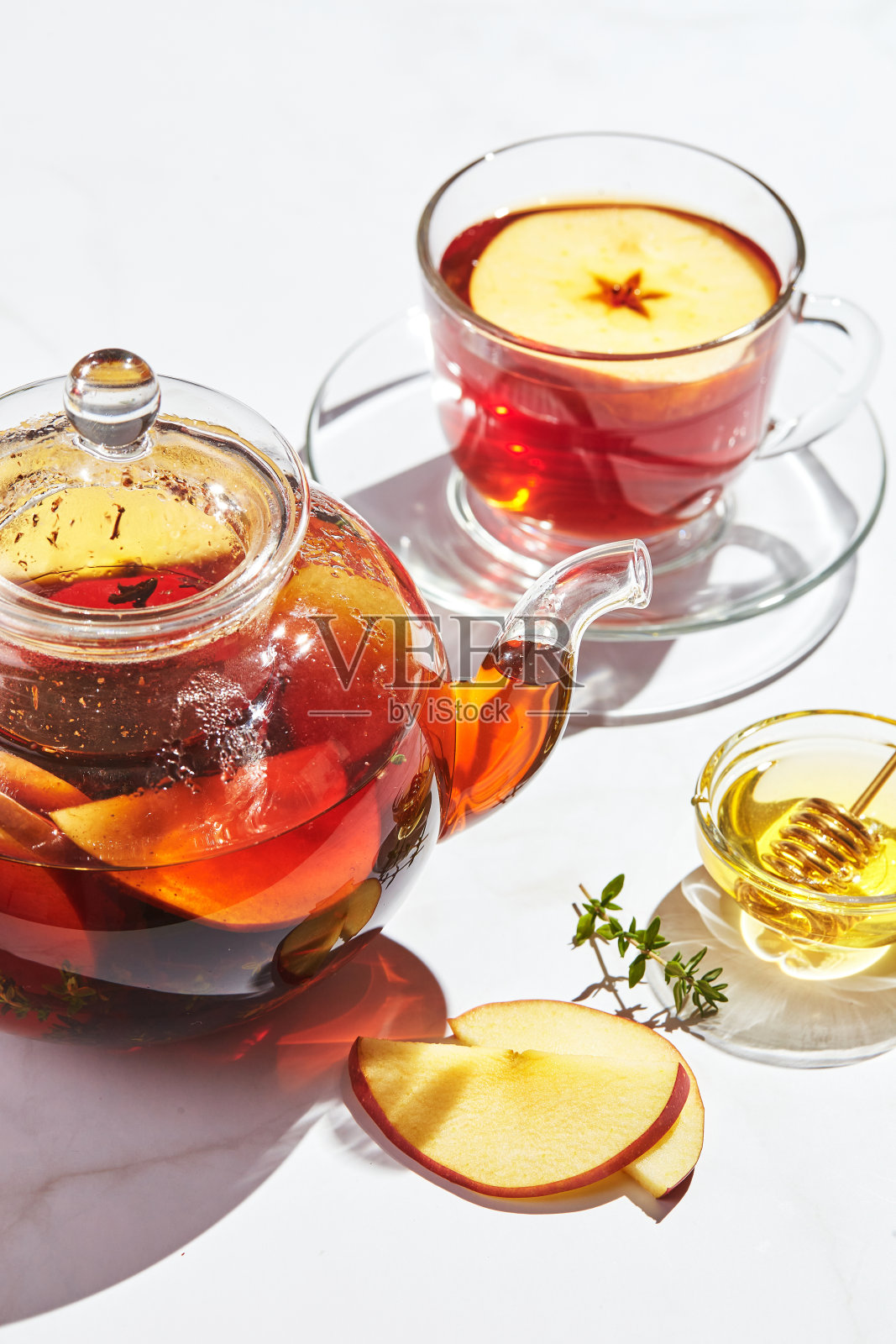 水果茶与苹果、百里香和蜂蜜在玻璃茶壶和杯子在白色背景与硬阴影照片摄影图片