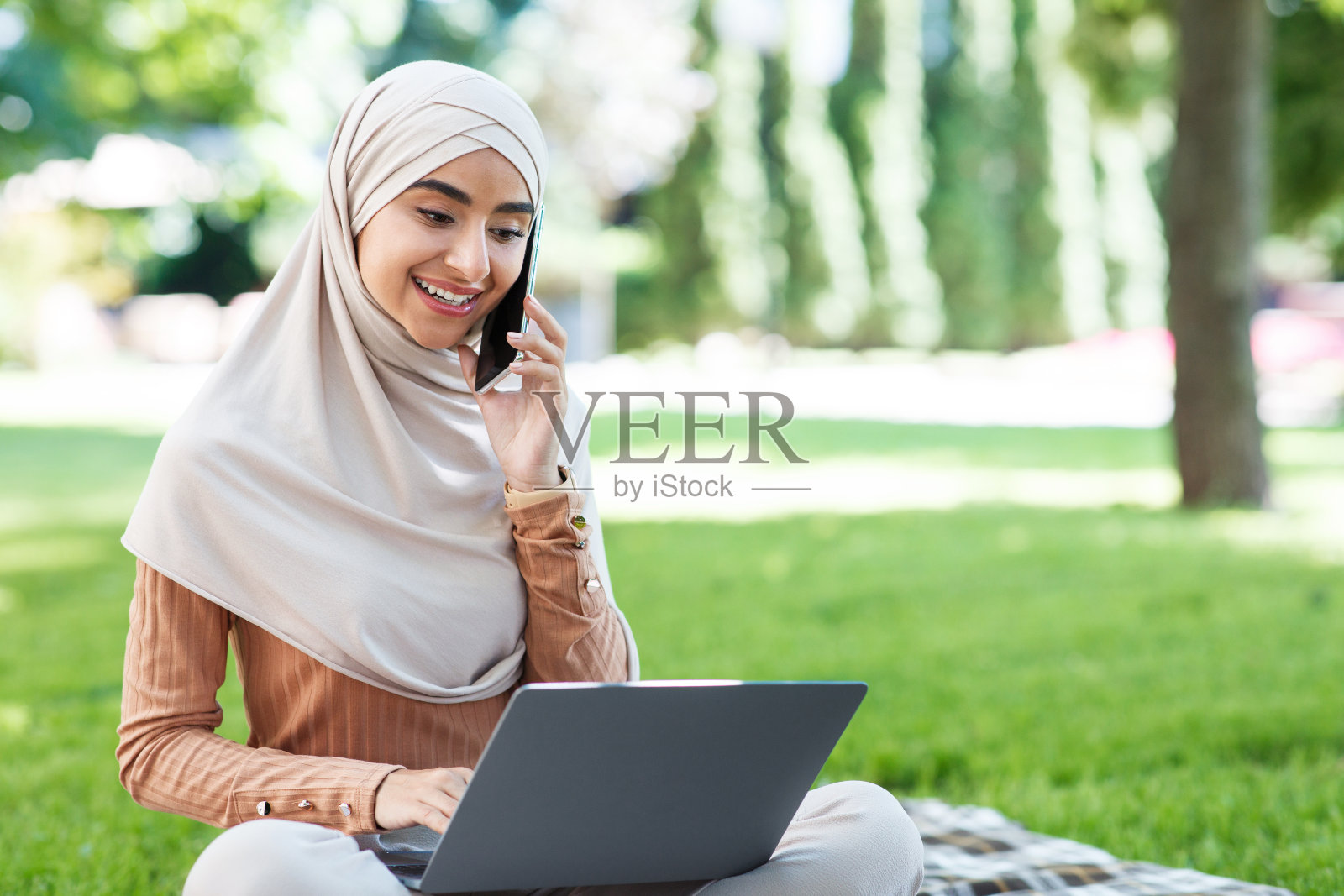 很高兴有吸引力的年轻中东女性戴着头巾看着笔记本电脑，打电话，在公园远程工作照片摄影图片