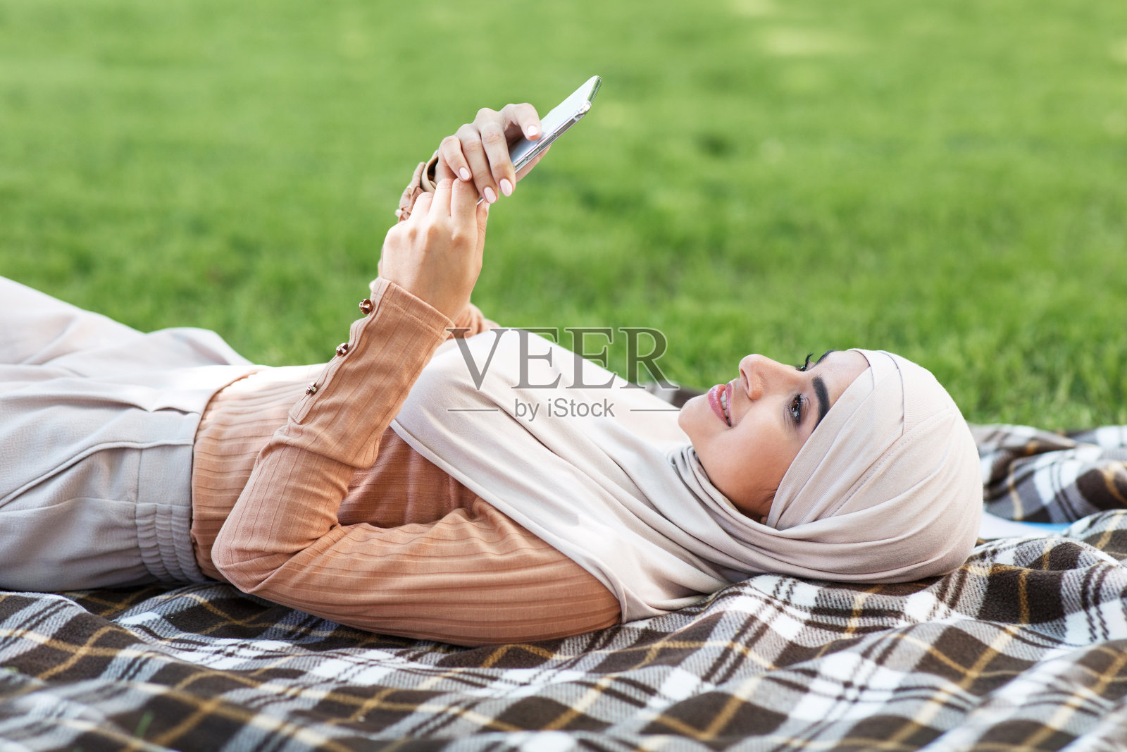 微笑迷人的年轻伊斯兰女士戴着头巾躺在格子花呢上，在公园里用手机看视频照片摄影图片