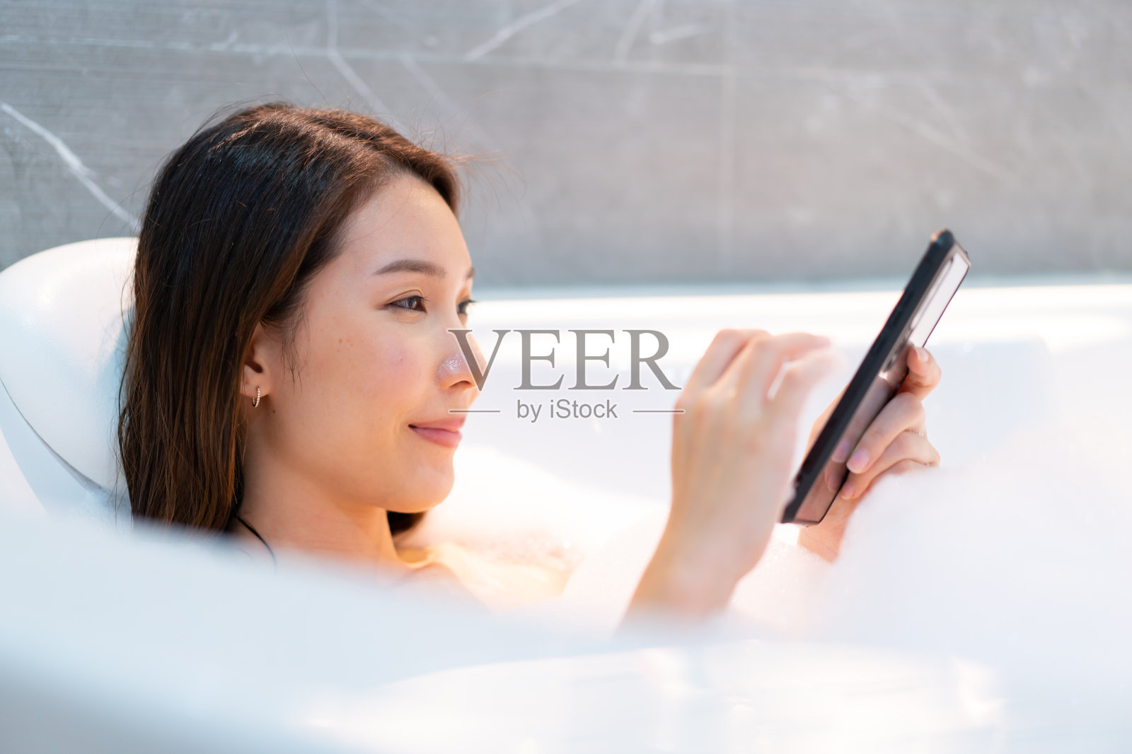 放松的亚洲女性使用智能手机洗澡泡沫浴缸。照片摄影图片