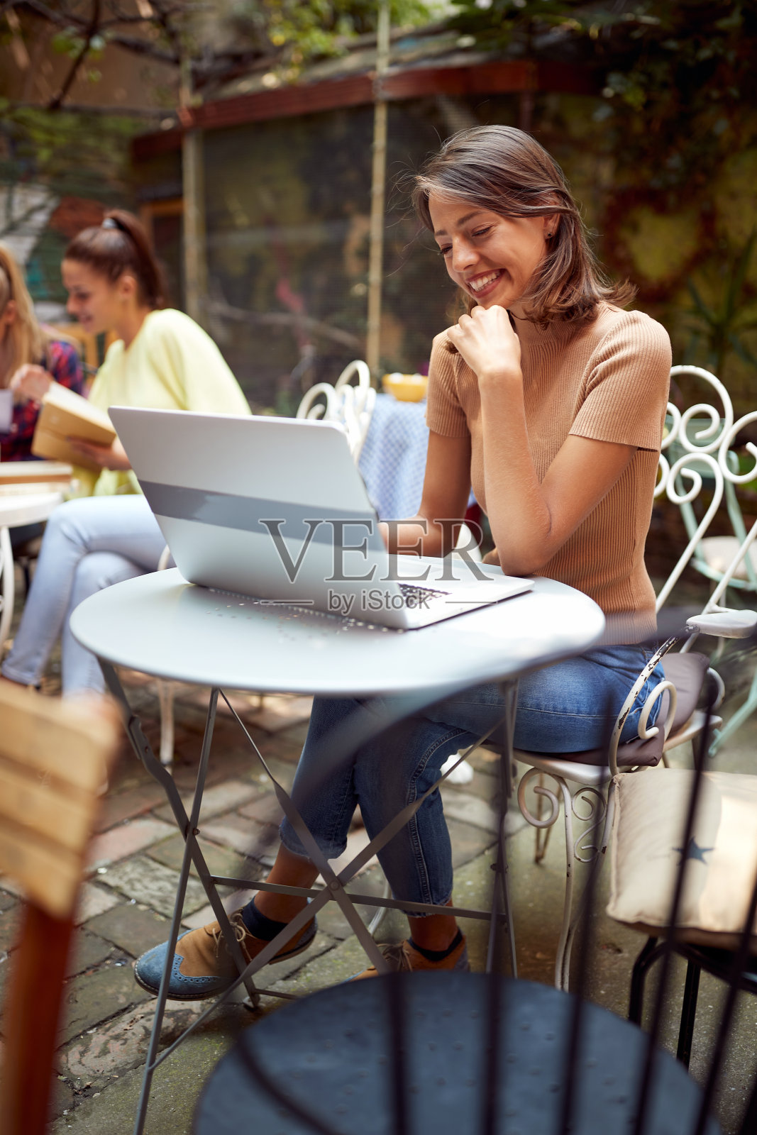 垂直图像的美丽的年轻女子坐在户外咖啡馆看着她的笔记本电脑在桌子上，微笑照片摄影图片