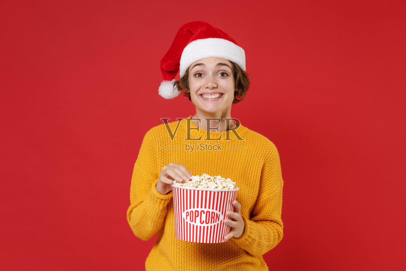 惊讶的年轻黑发圣诞老人女人穿着休闲的黄色毛衣圣诞帽拿着一桶爆米花孤立在红色背景的摄影棚肖像。新年庆祝快乐节日概念。照片摄影图片