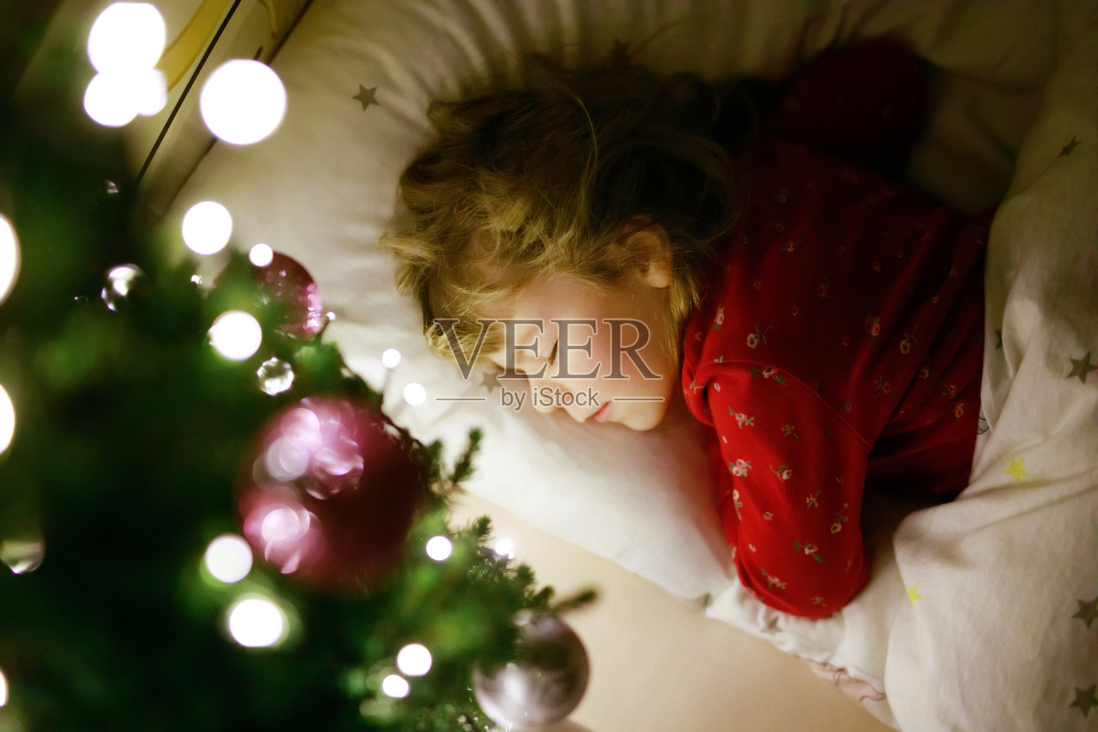 可爱的小女孩睡在圣诞树下，梦见圣诞老人在家里，在室内。传统的基督教节日。快乐的孩子等待圣诞节的礼物。温馨柔和的光照片摄影图片
