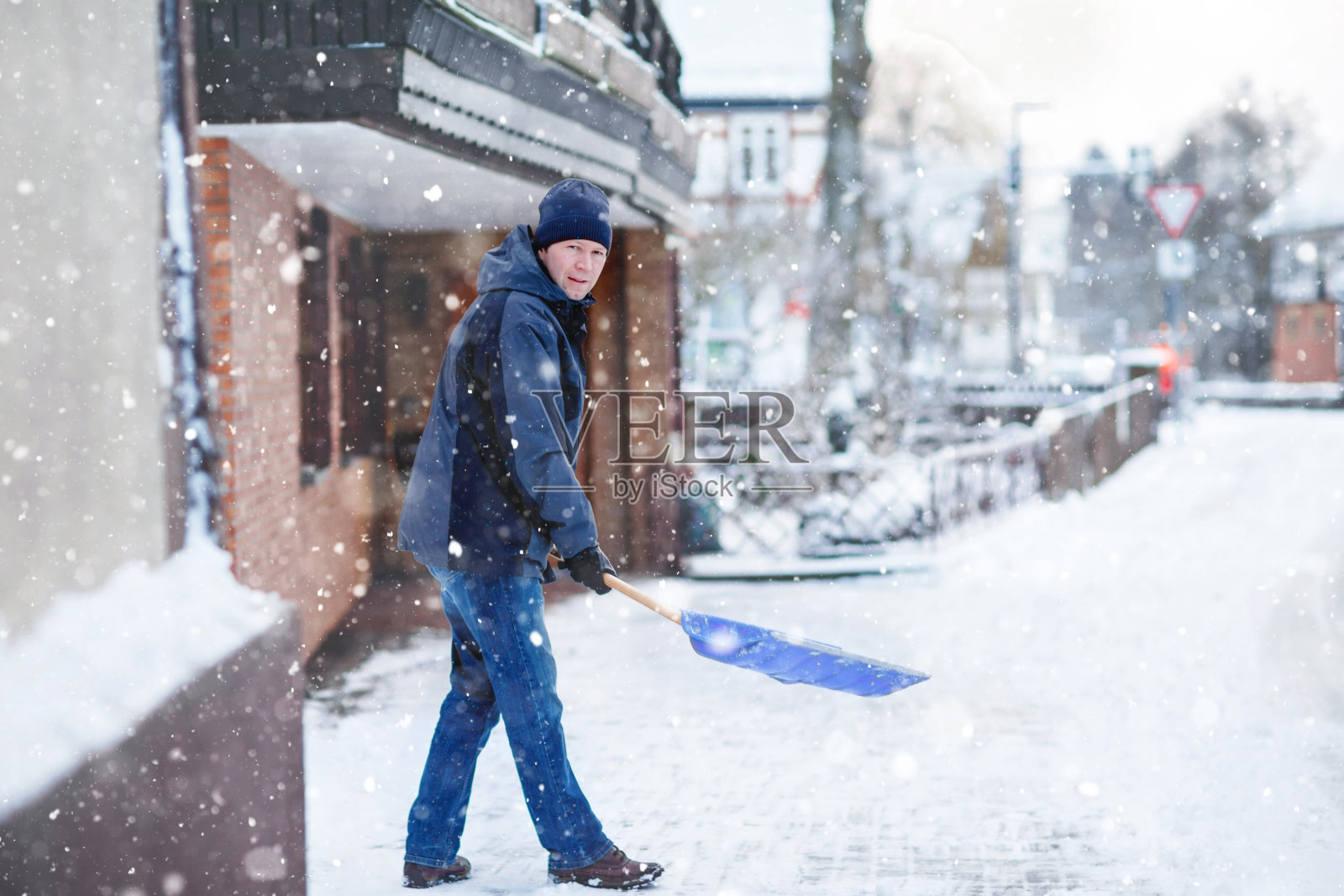 一名男子在冬天下雪时用雪铲清理人行道。欧洲的冬季。年轻人穿着温暖的冬装。德国降雪和天气混乱。暴风雪和大雪。Schneechaos照片摄影图片