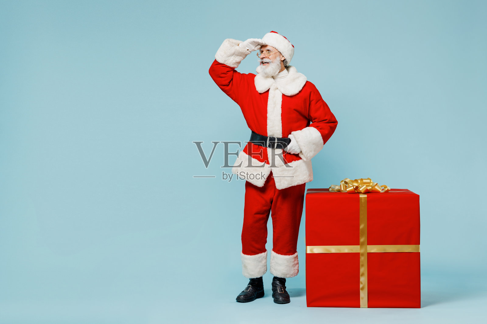 全身都是圣诞老人，戴着圣诞帽，穿着红色西装，站在大礼盒旁边，远远地孤立在纯蓝色背景的工作室里。2022年新年快乐庆典，圣诞快乐概念照片摄影图片