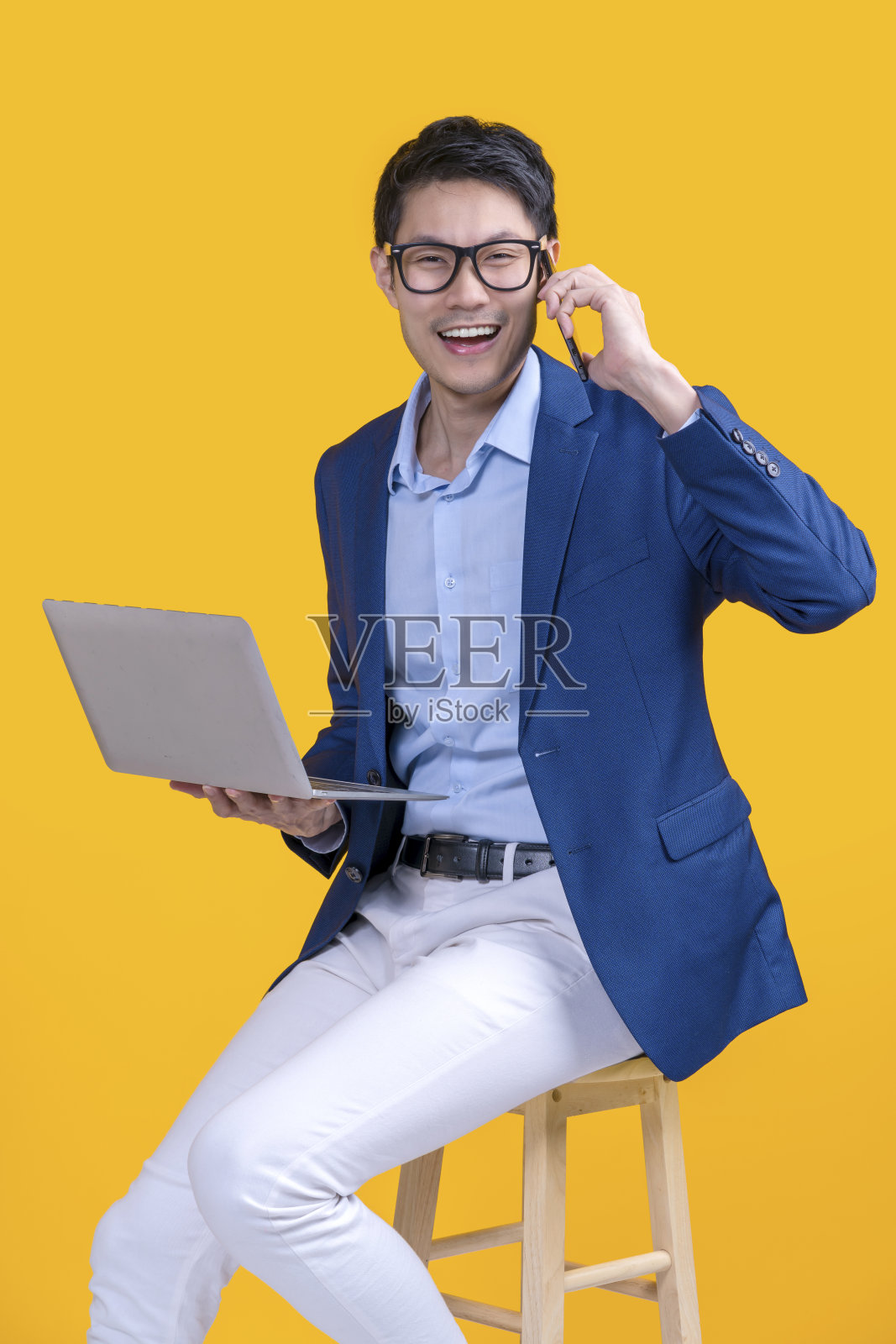 聪明的休闲眼镜，亚洲男性正装，坐在木凳上，手拿笔记本电脑，休闲放松，愉快微笑的工作，黄色背景的工作室拍摄，商务休闲的生活方式工作照片摄影图片